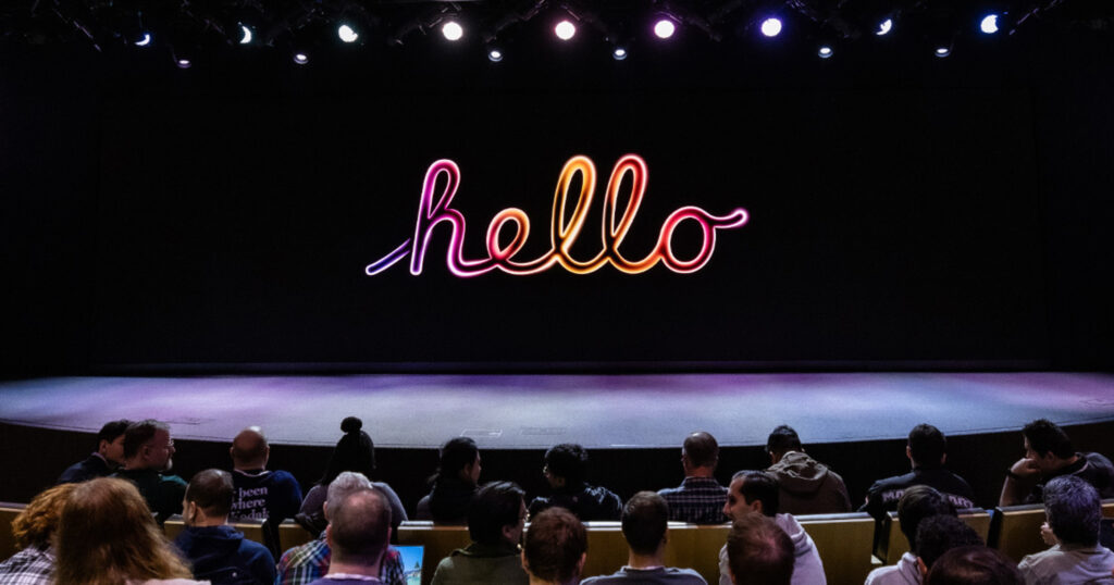 An audience sits in a dark auditorium looking at a large stage screen displaying the word "hello" in a glowing cursive font. The script features a vibrant gradient transition from purple and pink to orange and yellow. Stage lights shine from above, illuminating the scene and casting a soft glow on the audience.
