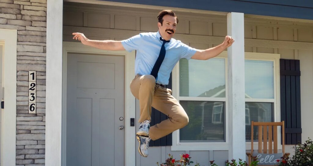 ‘Ted Lasso’ Season 4 Review: A Heartfelt Homecoming Jason Sudeikis wears a blue shirt and tie and jumps in front of a house.