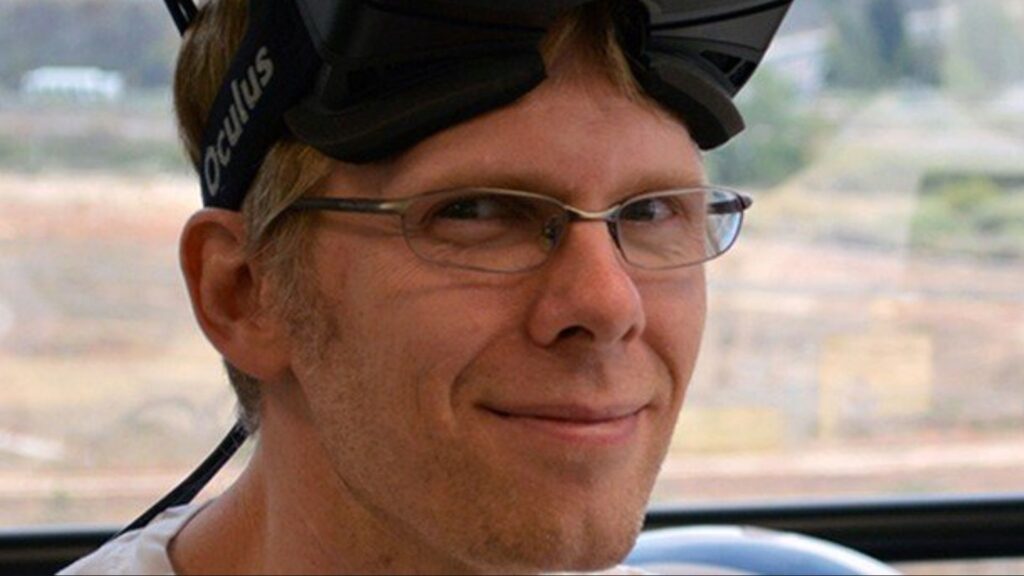 Programmer John Carmack smiling at camera with VR headset lifted over forehead in 2016.