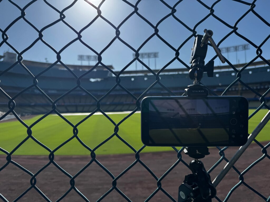 Apple’s MLB Dodgers Live Broadcast Was Shot With iPhone 18 Pro Max A phone mounted to a tripod behind a fence looking from the bullpen onto the field.