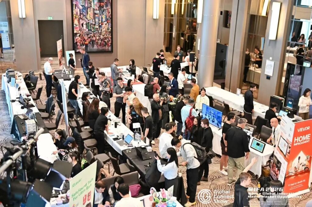 IFA-Nebenschau: Das haben wir auf der Global Connect Show getestet Vogelperspektive auf einen Hotelsaal mit mehreren Reihen von Ausstellungstischen, an denen Besucher und Standpersonal der Global Connect Show Technikprodukte zeigen und testen, im Hintergrund ein Werbebanner von Vevor.