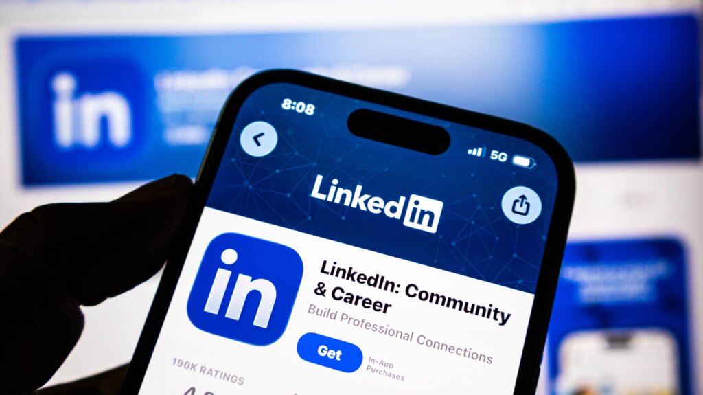 LinkedIn Adds More Profile Verifications to Combat AI-Powered Job Scams Closeup of a hand holding a smartphone displaying the linkedin app store page on a blurred background