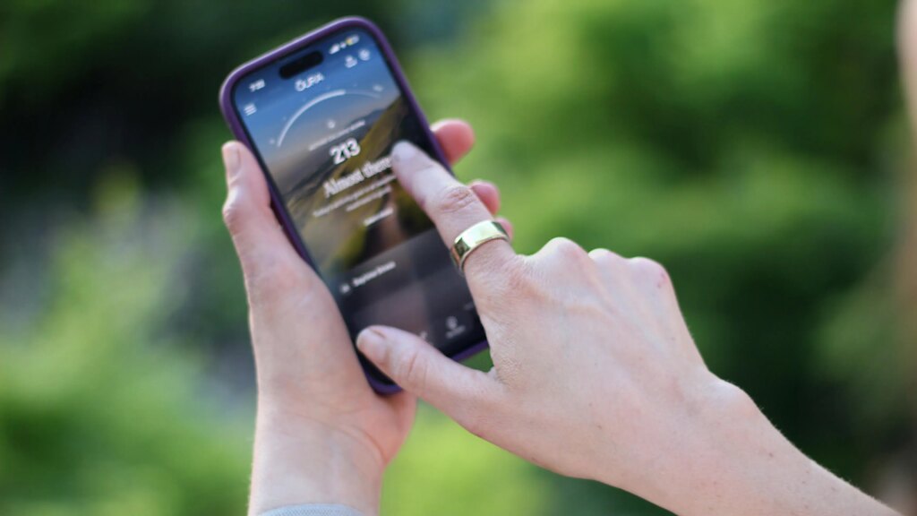 A finger wearing an Oura Ring pointing at a phone showing the Oura app.