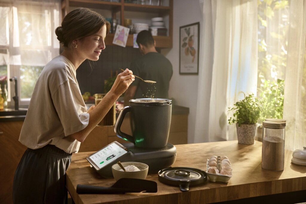 Eine Frau kocht in der Küche mit dem TM7 Thermomix von Vorwerk währen ihr Mann mit dem Rücken zu ihr steht.